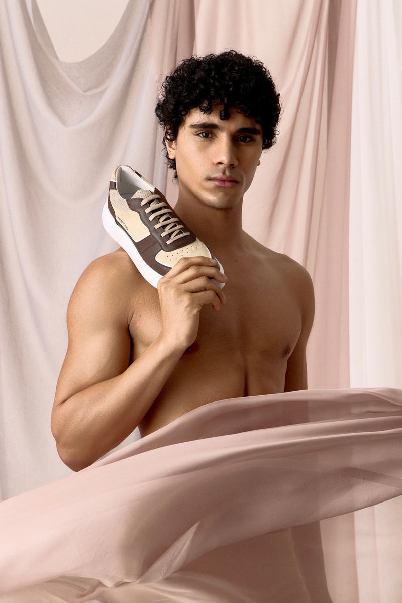 A shirtless adult male poses indoors with a brown and beige sneaker, draped with sheer fabric against a neutral backdrop.