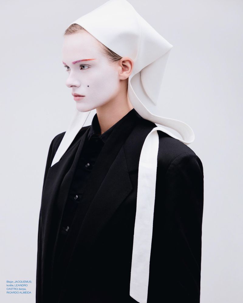 Model with white face makeup wearing a black blazer and white head covering against a pale background