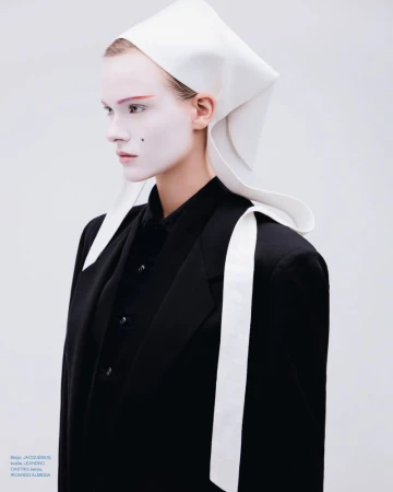 Model with white face makeup wearing a black blazer and white head covering against a pale background