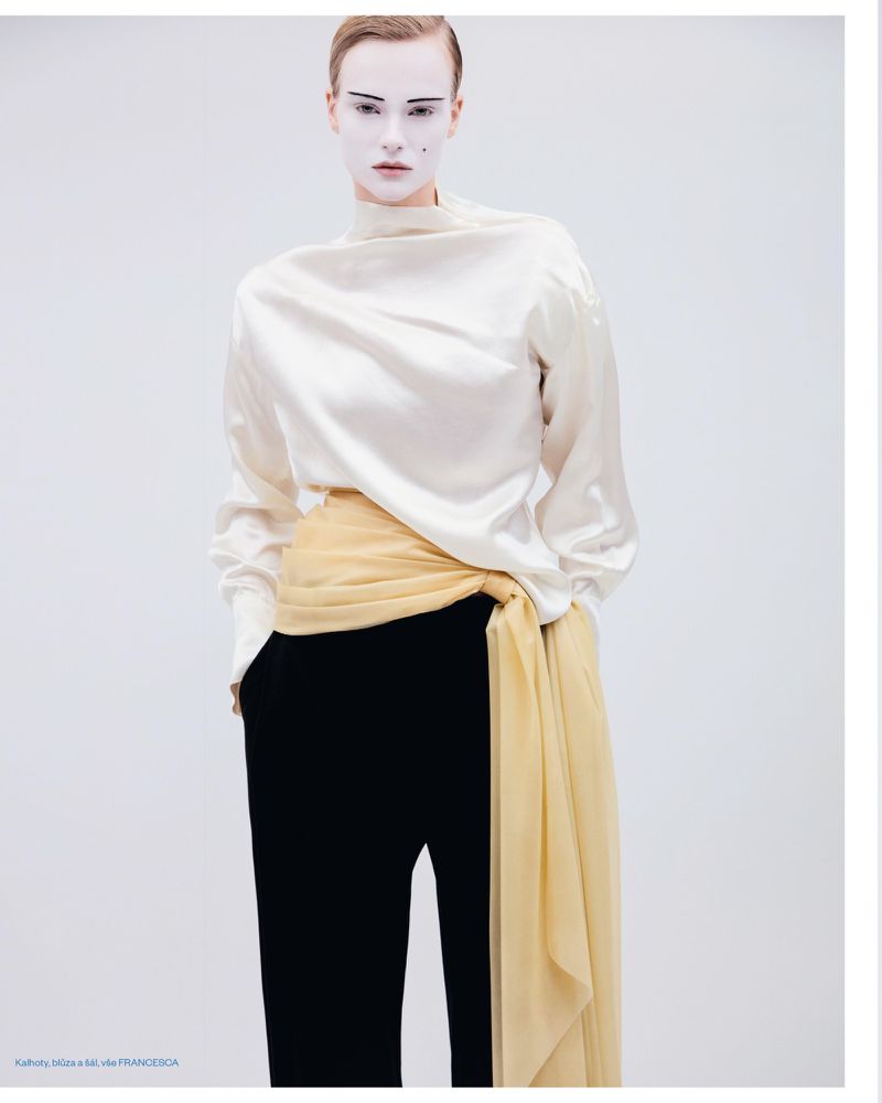 Model with white face makeup wearing a white silk top, black trousers, and a draped yellow sash against a seamless background