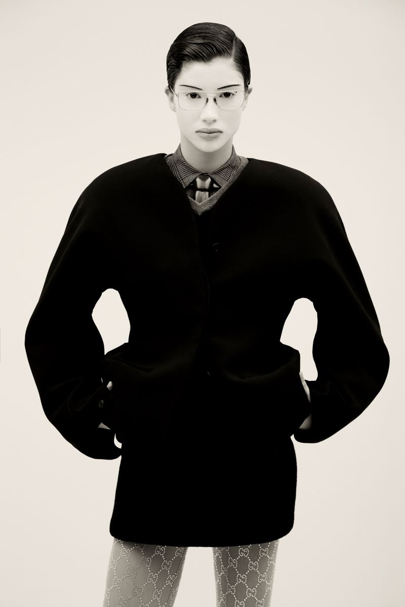 Model in a structured black coat wearing glasses and patterned tights against a light background