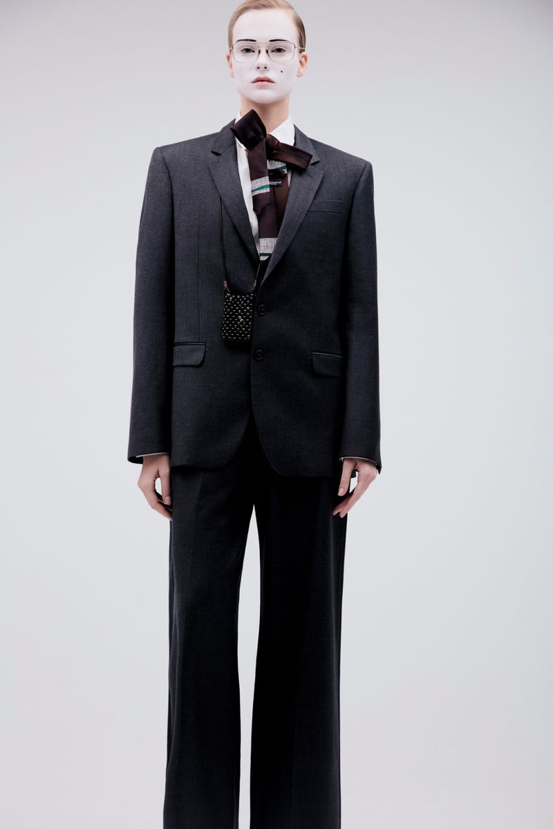 Model wearing a dark grey suit and tie with white face makeup against a seamless grey background