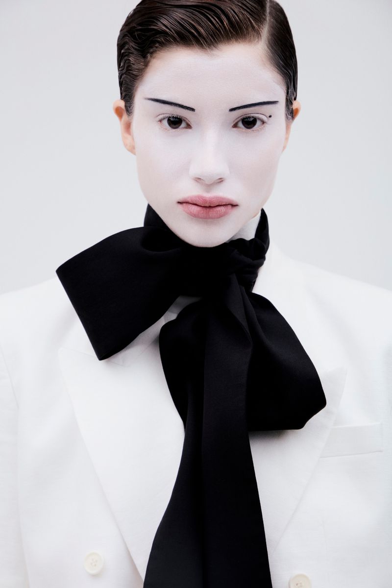 Close-up portrait of a model with pale white face makeup and thin eyebrows wearing a white blazer and large black bow tie against a white background.