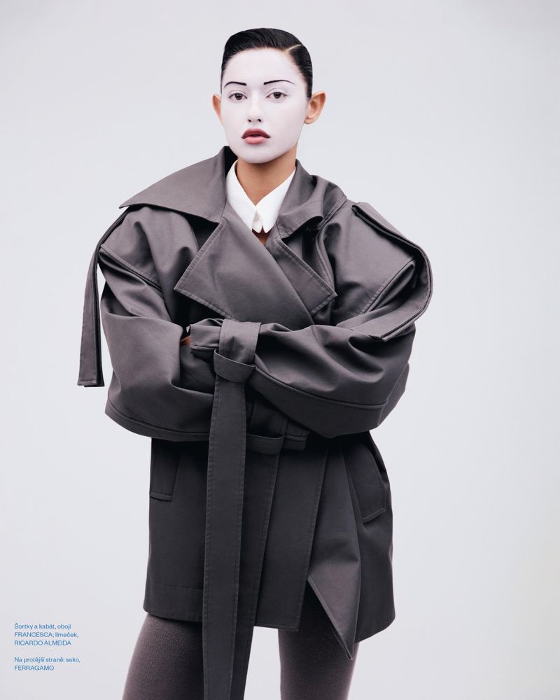 Fashion portrait of an adult woman wearing gray belted trench coat and pale foundation in a photo studio with a white backdrop.