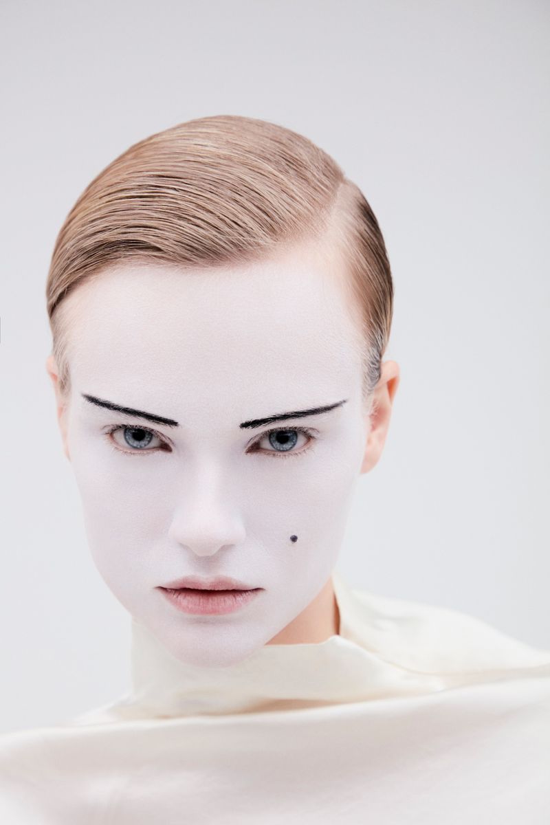 Close-up portrait of a model with white painted face and dark arched eyebrows against a light background.