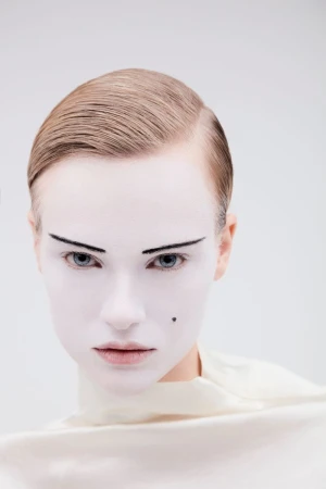 Close-up portrait of a model with white painted face and dark arched eyebrows against a light background.