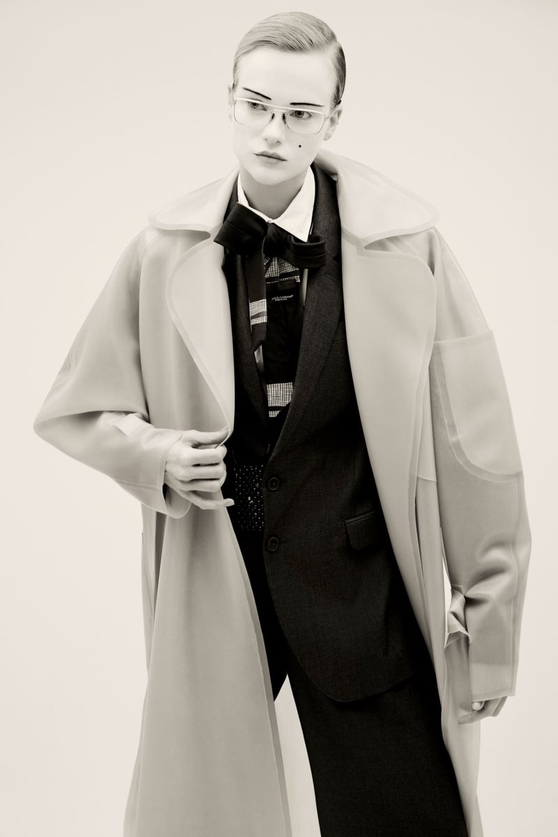 Model in tailored suit wearing oversized coat and eyeglasses against a plain background
