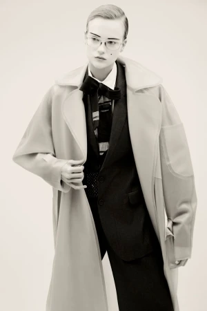 Model in tailored suit wearing oversized coat and eyeglasses against a plain background