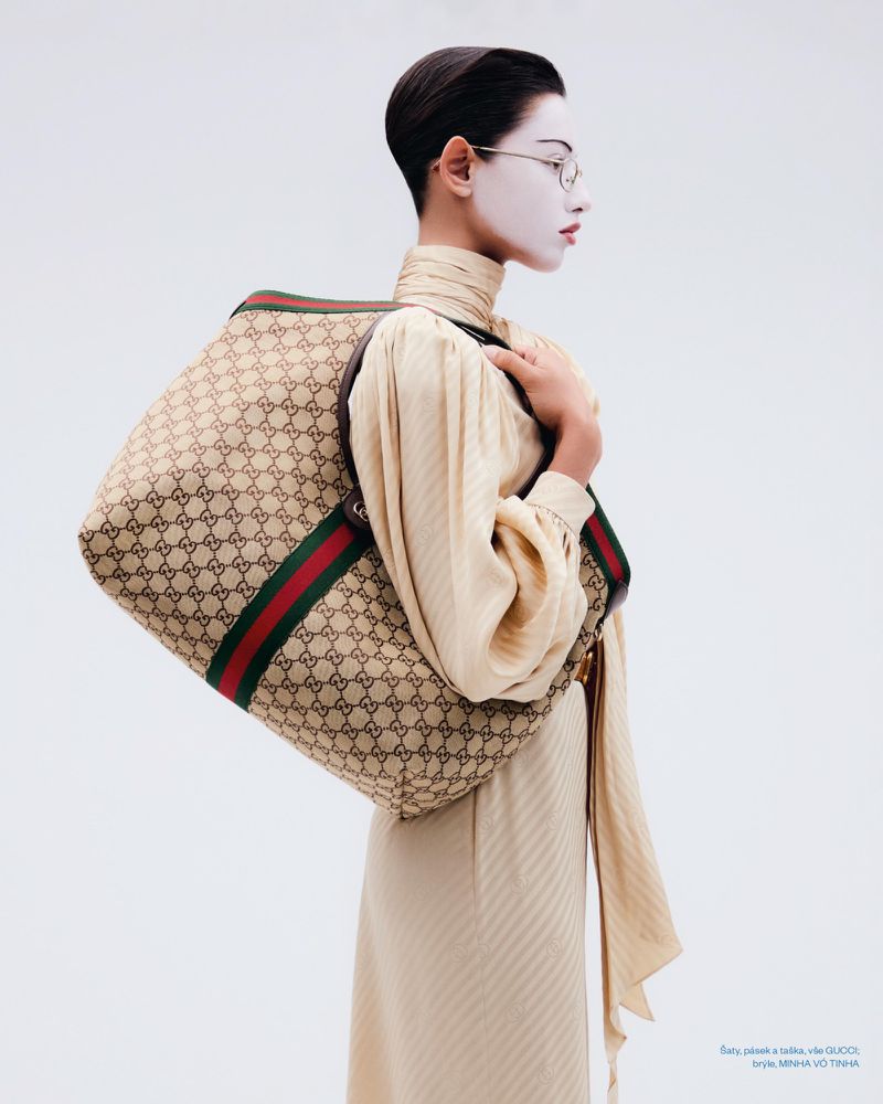 Model in profile wearing cream dress and eyeglasses holding a large patterned tote bag against white