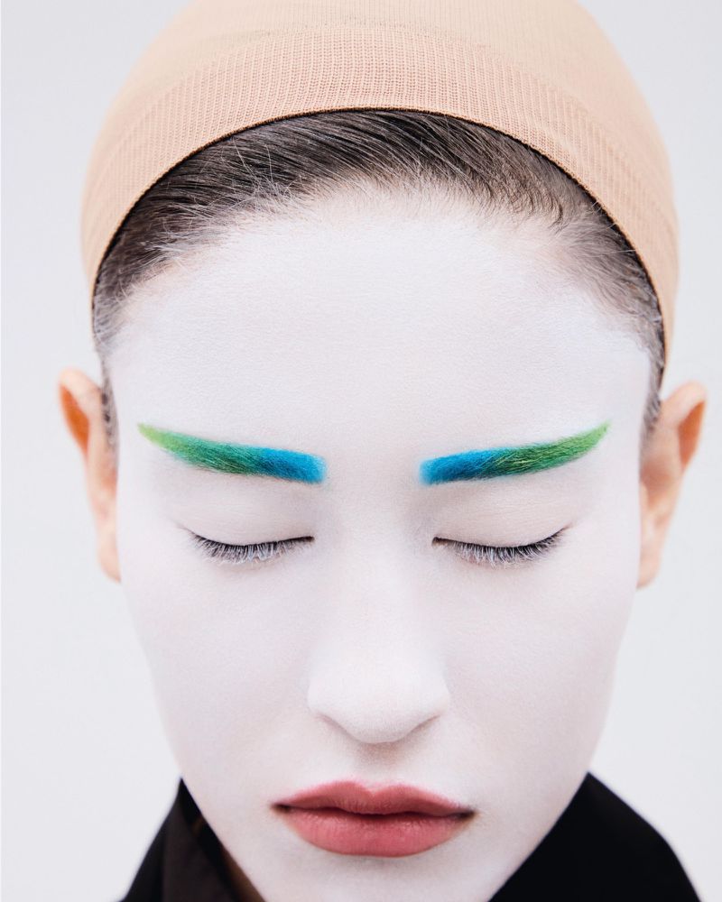 Close-up beauty portrait of a model with white face makeup, colorful blue and green eyebrows, and a nude wig cap against a plain white background.