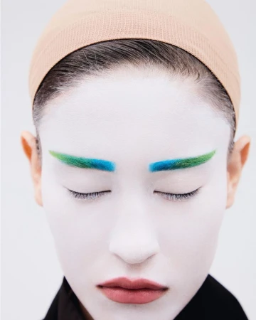 Close-up beauty portrait of a model with white face makeup, colorful blue and green eyebrows, and a nude wig cap against a plain white background.