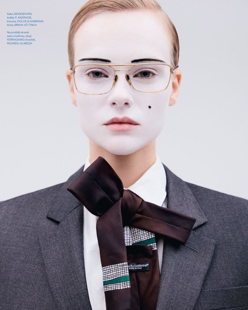 Portrait of a model with white face makeup wearing wire-rimmed glasses, a white collared shirt, dark tie, and a grey blazer.