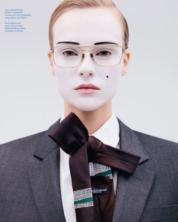 Portrait of a model with white face makeup wearing wire-rimmed glasses, a white collared shirt, dark tie, and a grey blazer.