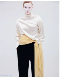 Model with white face makeup wearing a white silk top, black trousers, and a draped yellow sash against a seamless background
