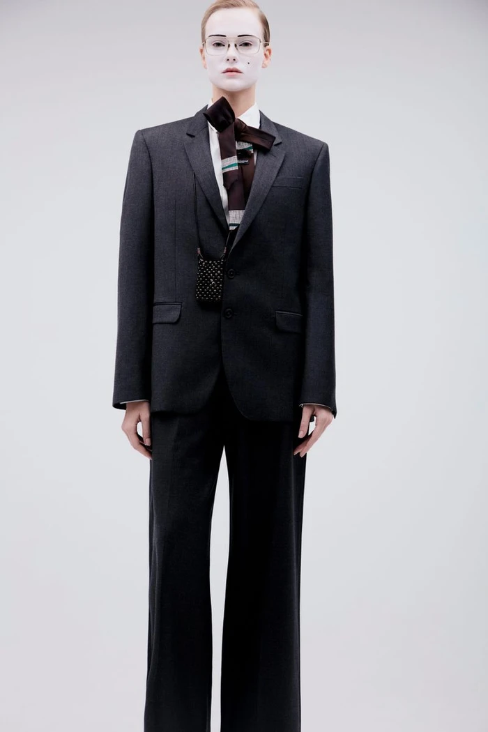 Model wearing a dark grey suit and tie with white face makeup against a seamless grey background