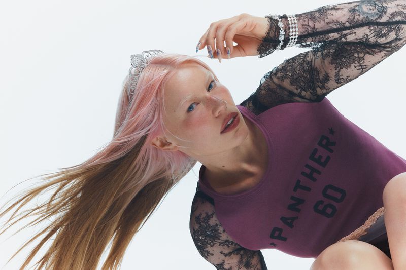 Model with pink and blonde hair wearing a tiara, purple top, and lace sleeves against a white background.