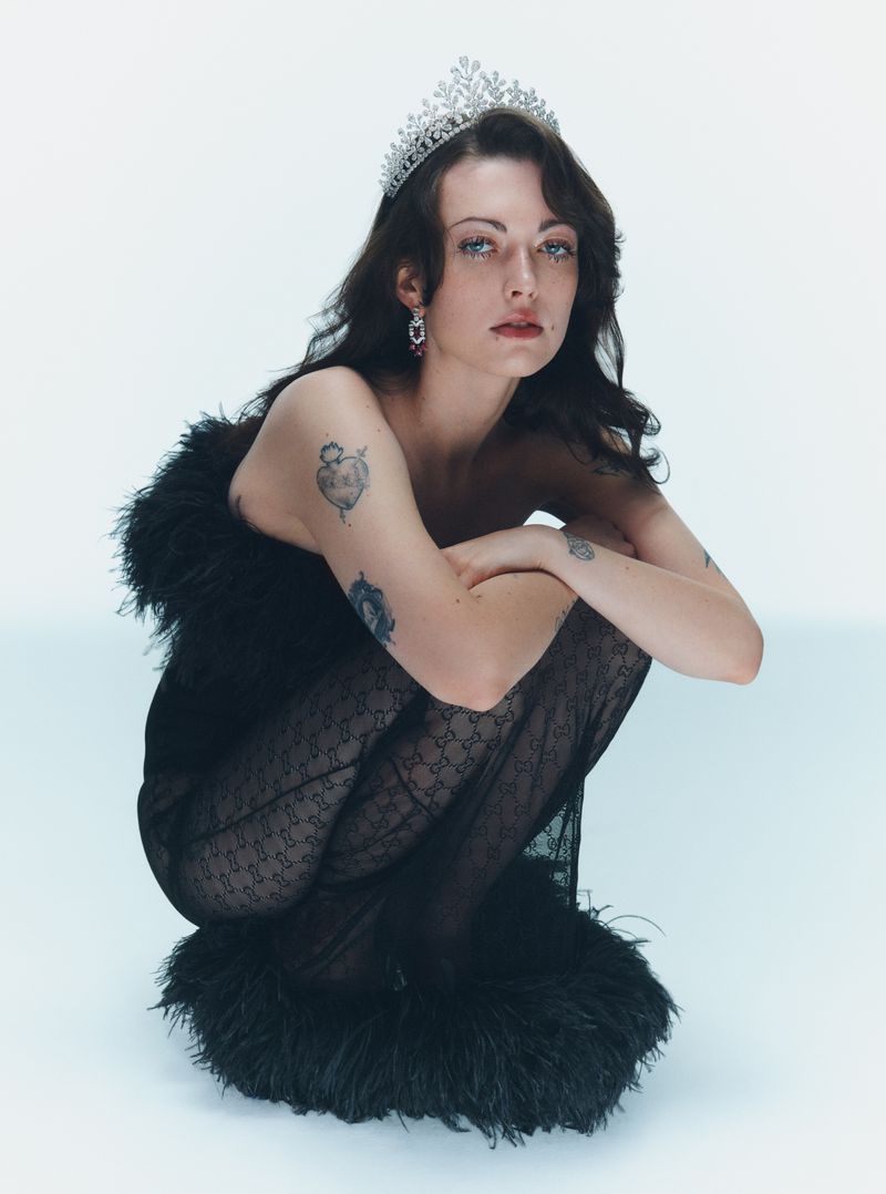 Woman wearing a tiara, black feather dress, and patterned tights crouching against a white background.