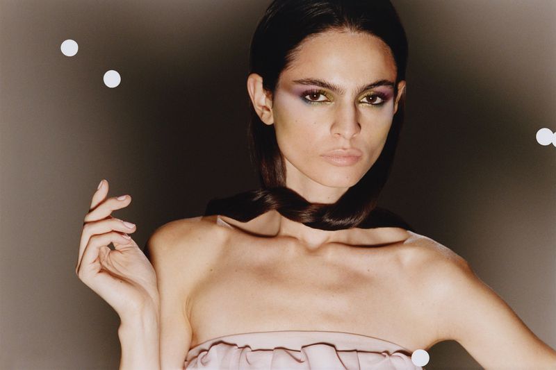 Close-up portrait of a female model with dramatic eye makeup and hair wrapped around her neck against a dark grey background.