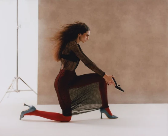 Model kneeling in mesh top and red tights against a studio backdrop holding a shoe