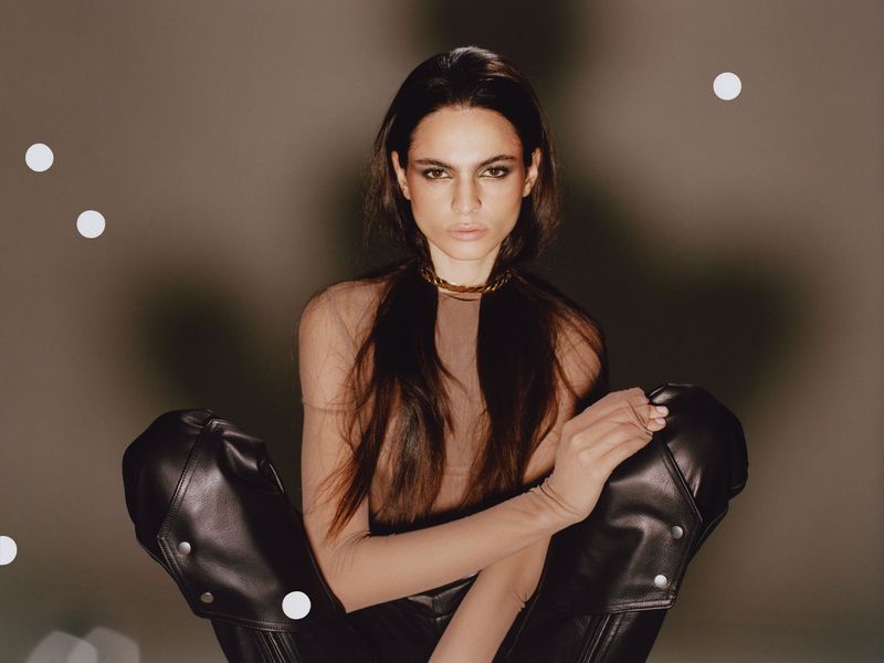 Model with long dark hair wearing a neutral top and black leather trousers against a muted grey studio background with soft bokeh lights.