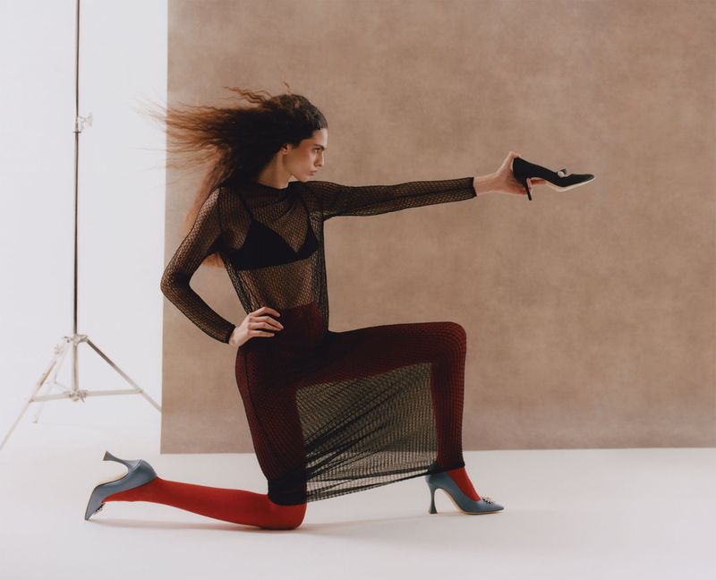 Model in sheer black mesh top holding a high-heeled shoe while kneeling against a neutral studio backdrop.