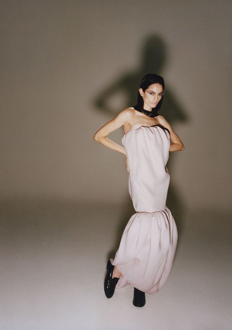 Model in pink sculptural dress and black shoes posing against a plain grey studio backdrop with a dramatic shadow cast
