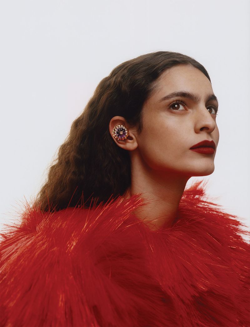 Portrait of a model wearing a textured red garment and ornate jewelled ear cuff against a plain white background