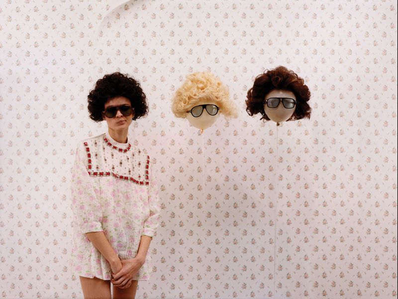 Model in a floral dress and dark sunglasses standing beside two floating balloon-like heads wearing wigs and glasses.