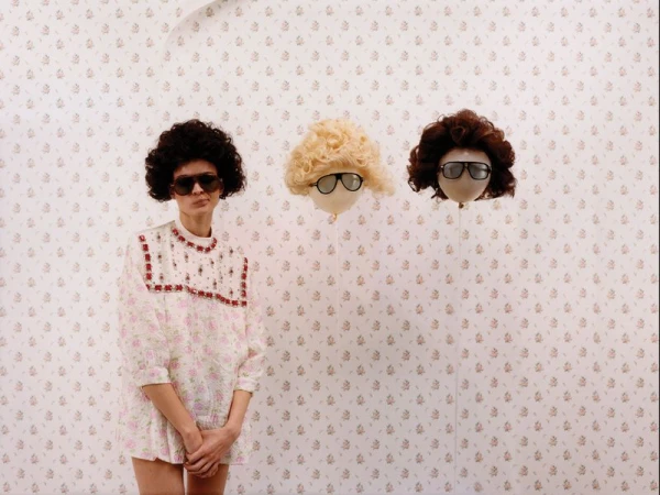 Model in a floral dress and dark sunglasses standing beside two floating balloon-like heads wearing wigs and glasses.