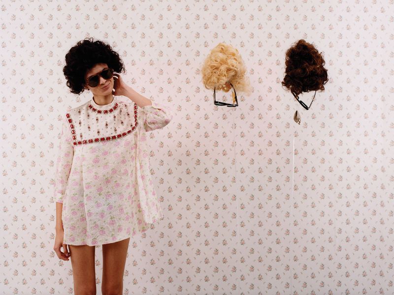 Model in floral dress with dark sunglasses standing next to floating wigs on wallpaper