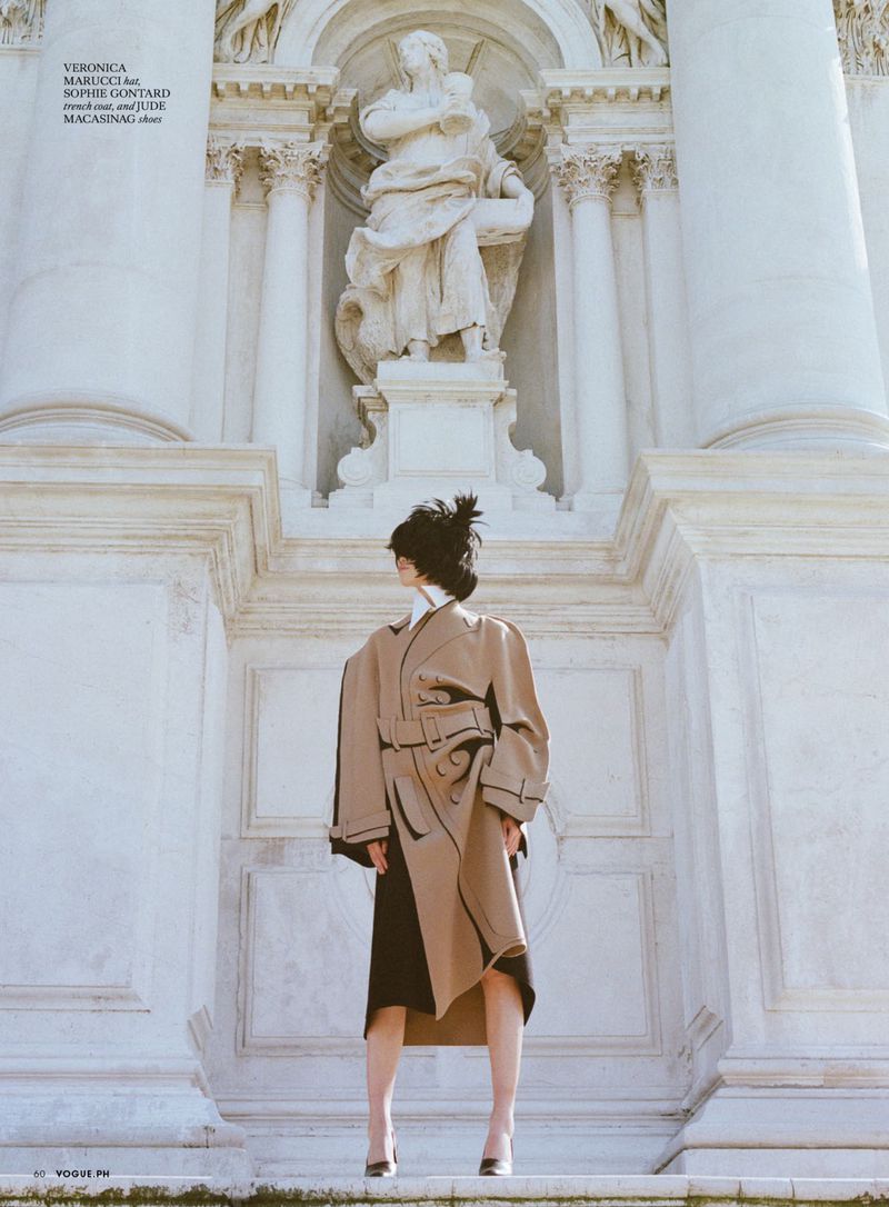 Model wearing tan structured coat standing before classic architecture and a statue for Vogue Philippines