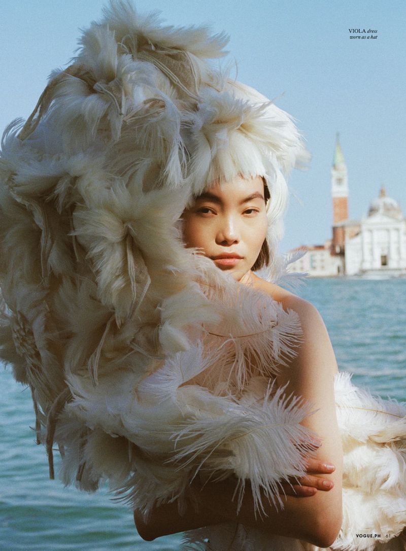 Model Maggie Cheng wearing a large white feathered hat against a waterfront backdrop with distant architecture for Vogue Philippines.