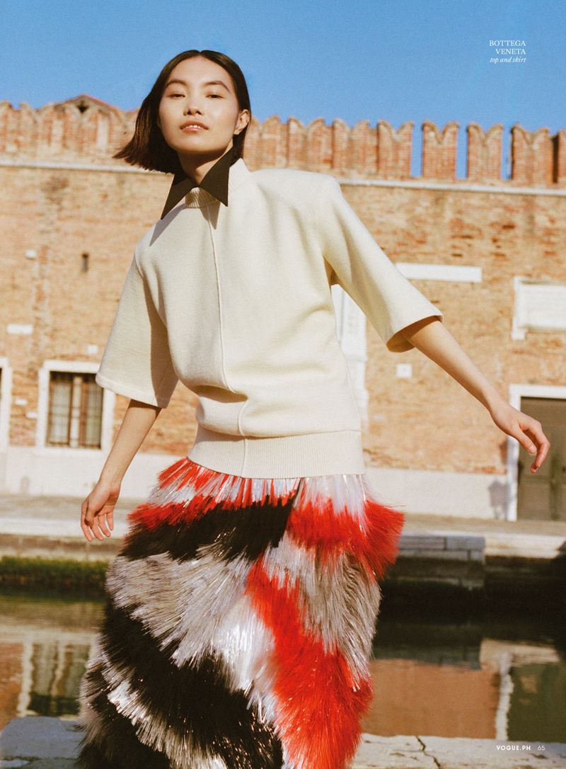 Model in a cream top and patterned skirt posing against a brick building facade and water for Vogue Philippines