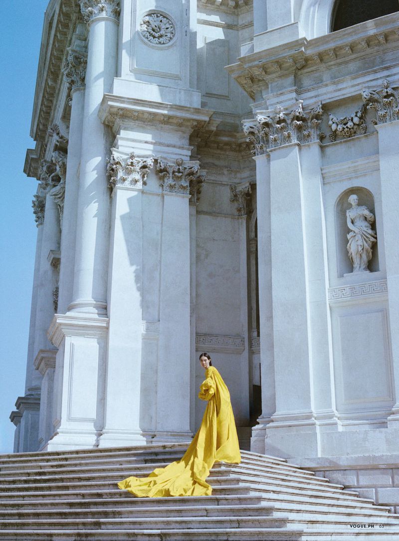 Model in a long yellow dress standing on grand stone steps before a monumental classical white building exterior.