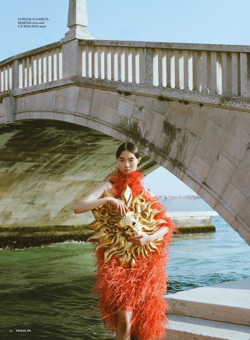 Model in vibrant red feather dress holding a golden sun mask beneath a stone bridge by water for Vogue Philippines La Vita editorial