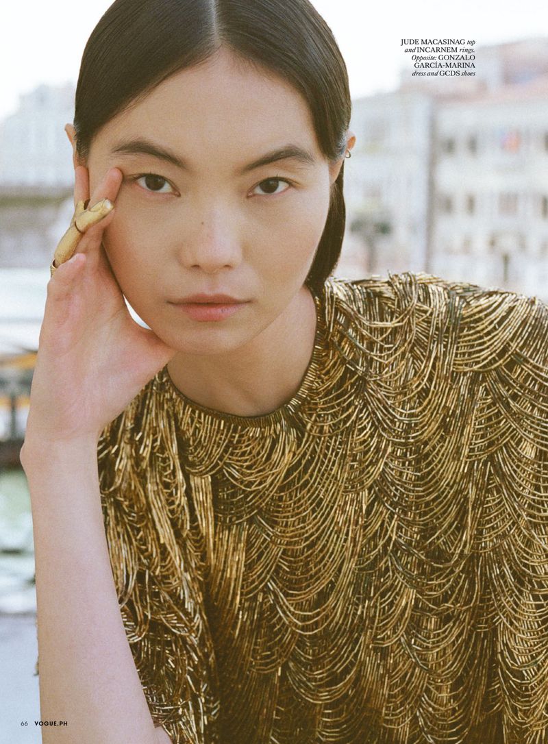 Close-up portrait of an East Asian model wearing a metallic gold beaded top with a gold ring on her hand, outdoors with a blurred background.