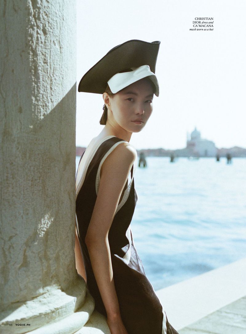 Model leaning against a stone column by the water wearing a dark dress and a structured hat for Vogue Philippines