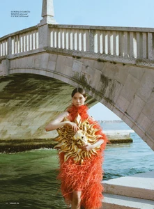 Model in vibrant red feather dress holding a golden sun mask beneath a stone bridge by water for Vogue Philippines La Vita editorial