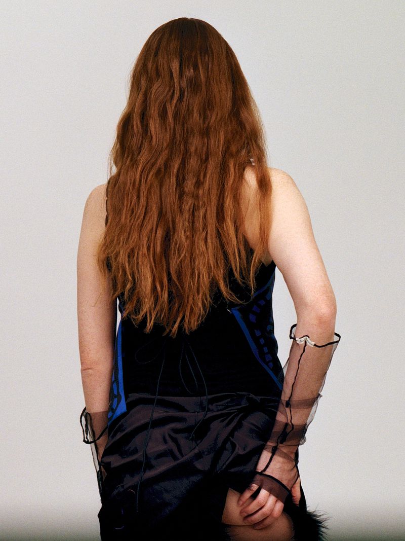 A woman with long wavy auburn hair is seen from the back, wearing a black and blue top with sheer gloves and a dark skirt.