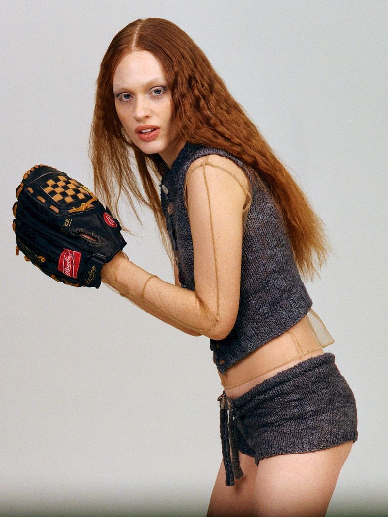 A slender woman with long auburn hair poses indoors wearing knit shorts, a vest, and a baseball glove against a neutral backdrop.
