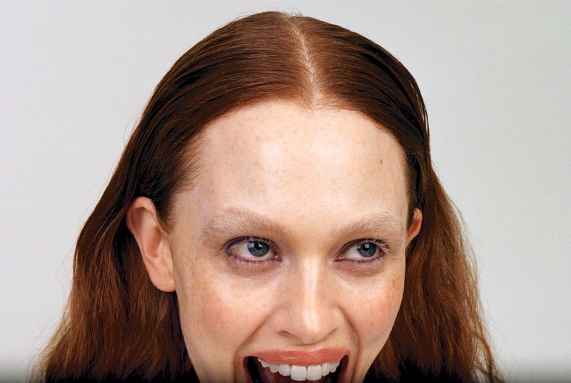 Close-up portrait of a fair-skinned adult female with auburn hair and light eyebrows, smiling widely in a brightly lit, neutral setting.