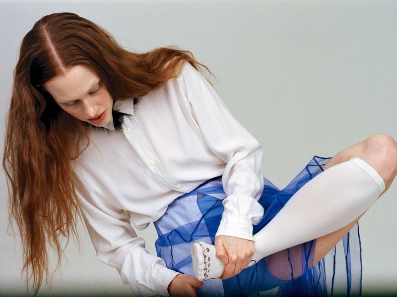 A fair-skinned woman with auburn hair poses in a white blouse and blue tulle skirt, seated with one leg bent and her hand on her ankle.