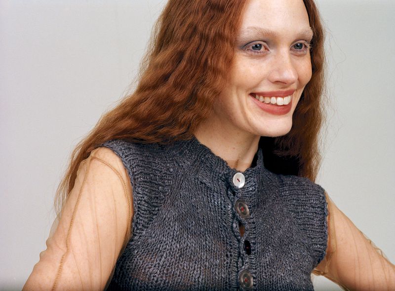 Smiling woman with auburn hair and wearing a grey, knitted vest, posing in front of a neutral background for a fashion portrait.