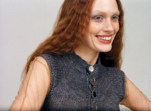 Smiling woman with auburn hair and wearing a grey, knitted vest, posing in front of a neutral background for a fashion portrait.