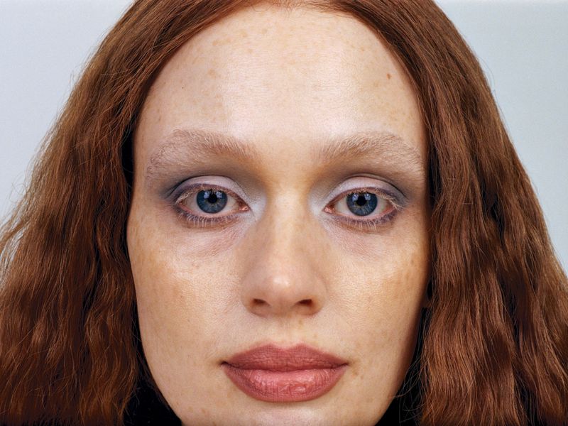 Close-up portrait of a red-haired woman with blue eyes, fair skin, and freckles, showcasing beauty and makeup details on a light grey background.