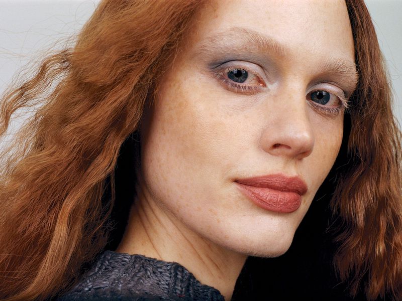 Close-up portrait of a redhead woman with fair skin, freckles, and smokey eye makeup. She wears a dark sweater against a neutral background.