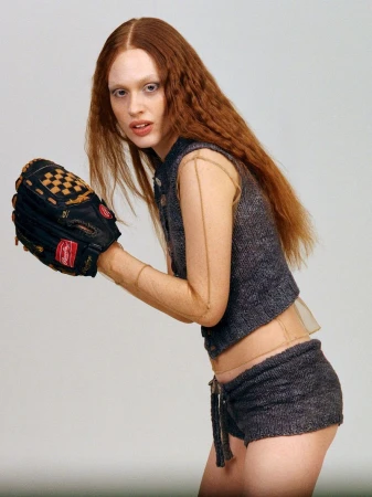 Model Laura Schuller with crimped auburn hair wearing a grey knit vest and matching shorts while holding a baseball glove.