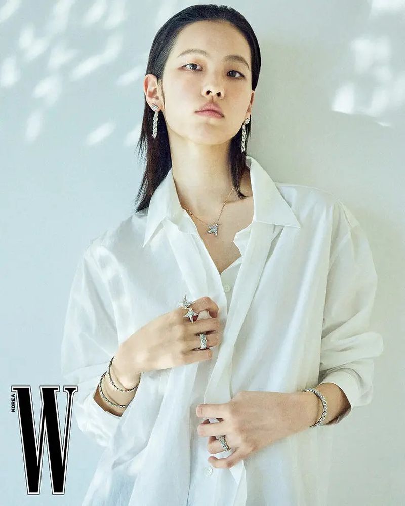 Model wearing a white button-down shirt and silver jewelry against a pale wall with dappled light shadows.