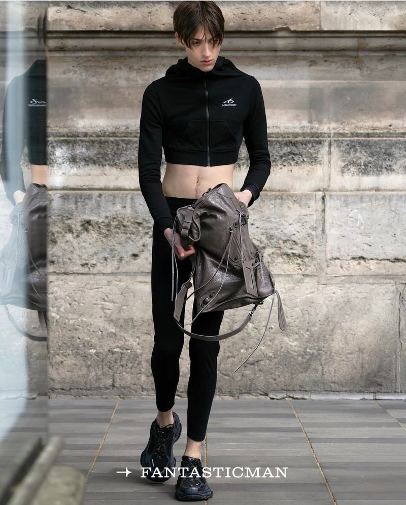 Model wearing a black crop hoodie and dark trousers holding a large leather bag outdoors