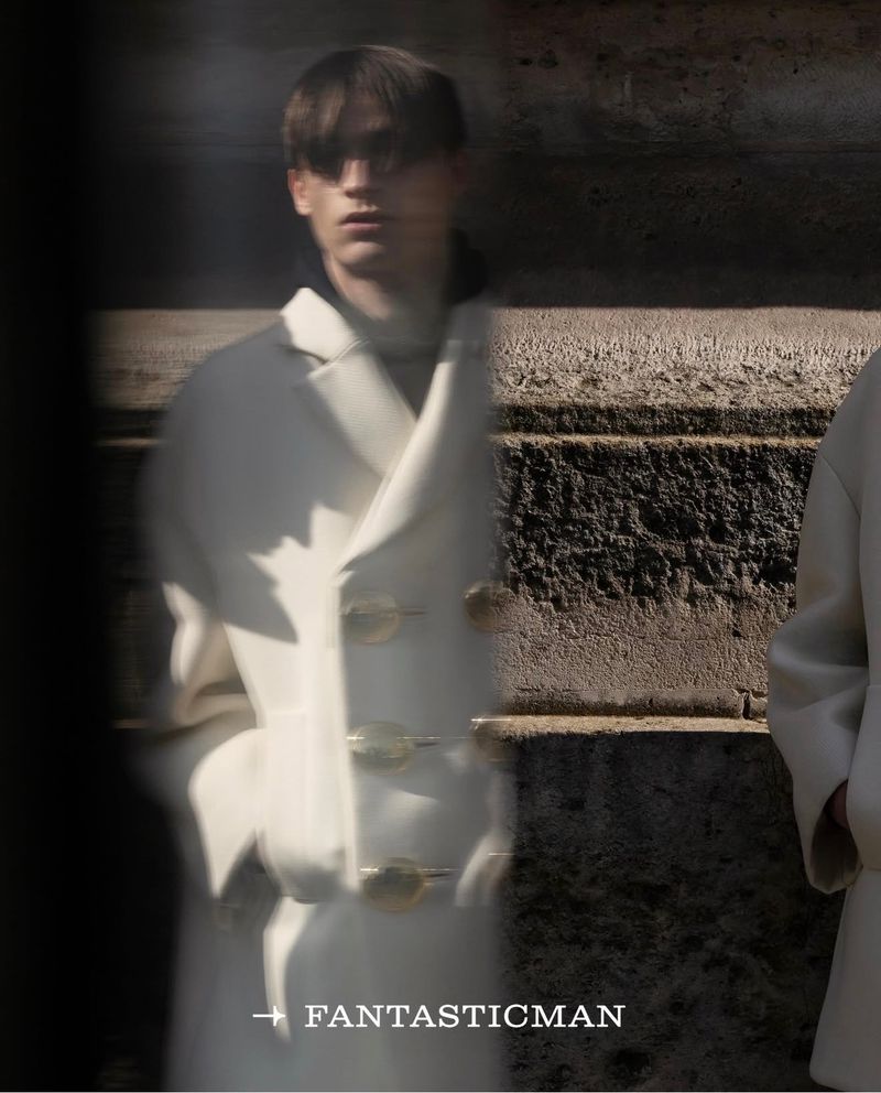 Indoor fashion shot featuring a blurred male model in a cream-colored double-breasted coat with metallic closures against a stone wall.